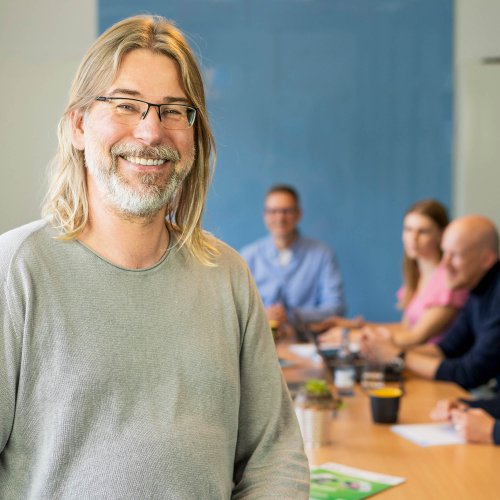 DVZ Mitarbeiter Uwe steht im Vordergrund, im Hintergrund halten DVZ-Mitarbeiter*innen ein Meeting ab