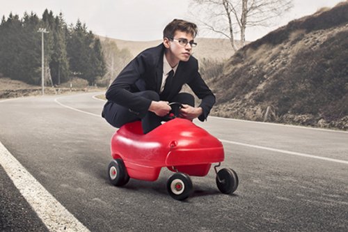 Abbildung: Ein junger Mann mit Anzug und Brille fährt in einem Bobby Car eine Straße entlang.